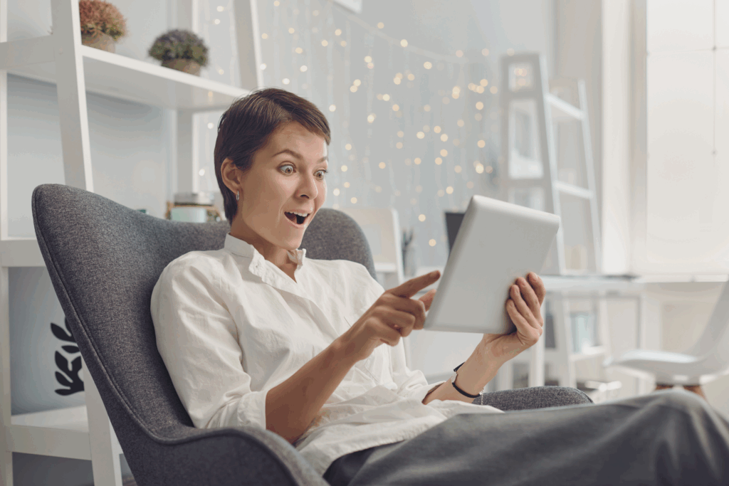 A lady sitting on the couch looking at an iPad with a shocked look on her face.